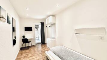 Ein modernes, helles Schlafzimmer mit Bett, Schreibtisch und Stuhl. Ein Kleiderschrank mit Platz zum Aufhängen und ein kleines Fenster mit Vorhängen.
