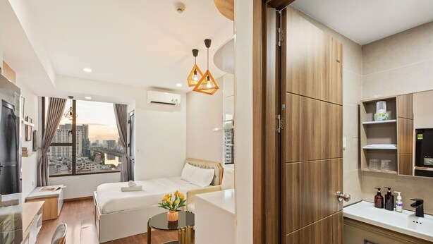 A modern hotel room with a bed, a desk, a chair, a small table with flowers, a view of the city, and a bathroom with a mirror and shelves. - SaiGon Rivergate (Ho Chi Minh City)