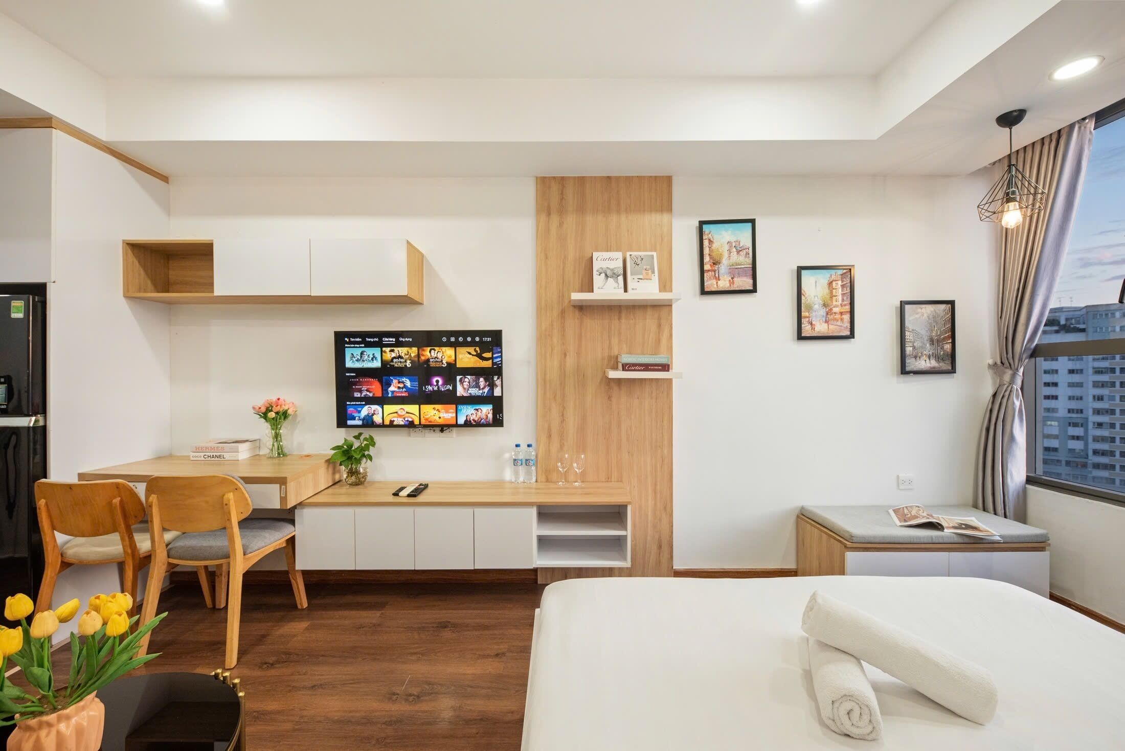 Ein modernes Hotelzimmer mit einem Holztisch, einem an der Wand befestigten Fernseher, einem Bett mit einem zusammengerollten Handtuch und einem Fenster mit Vorhängen.