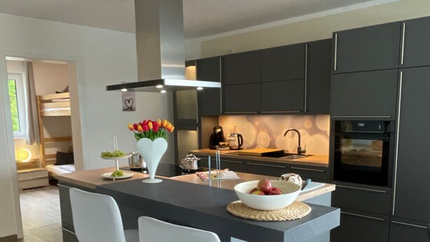 A modern kitchen with a central island, two white bar stools, dark cabinetry, and a built-in oven. - Apartment 6 "Petra" (Sankt Andreasberg)
