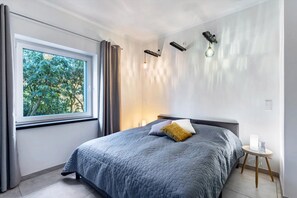 Un dormitorio con cama, ventana con cortinas, mesita de noche con lámpara e iluminación en la pared.