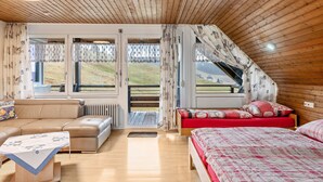 A cozy room with a wooden ceiling, a sofa, a bed with red bedding, and large windows offering a view of a grassy area.