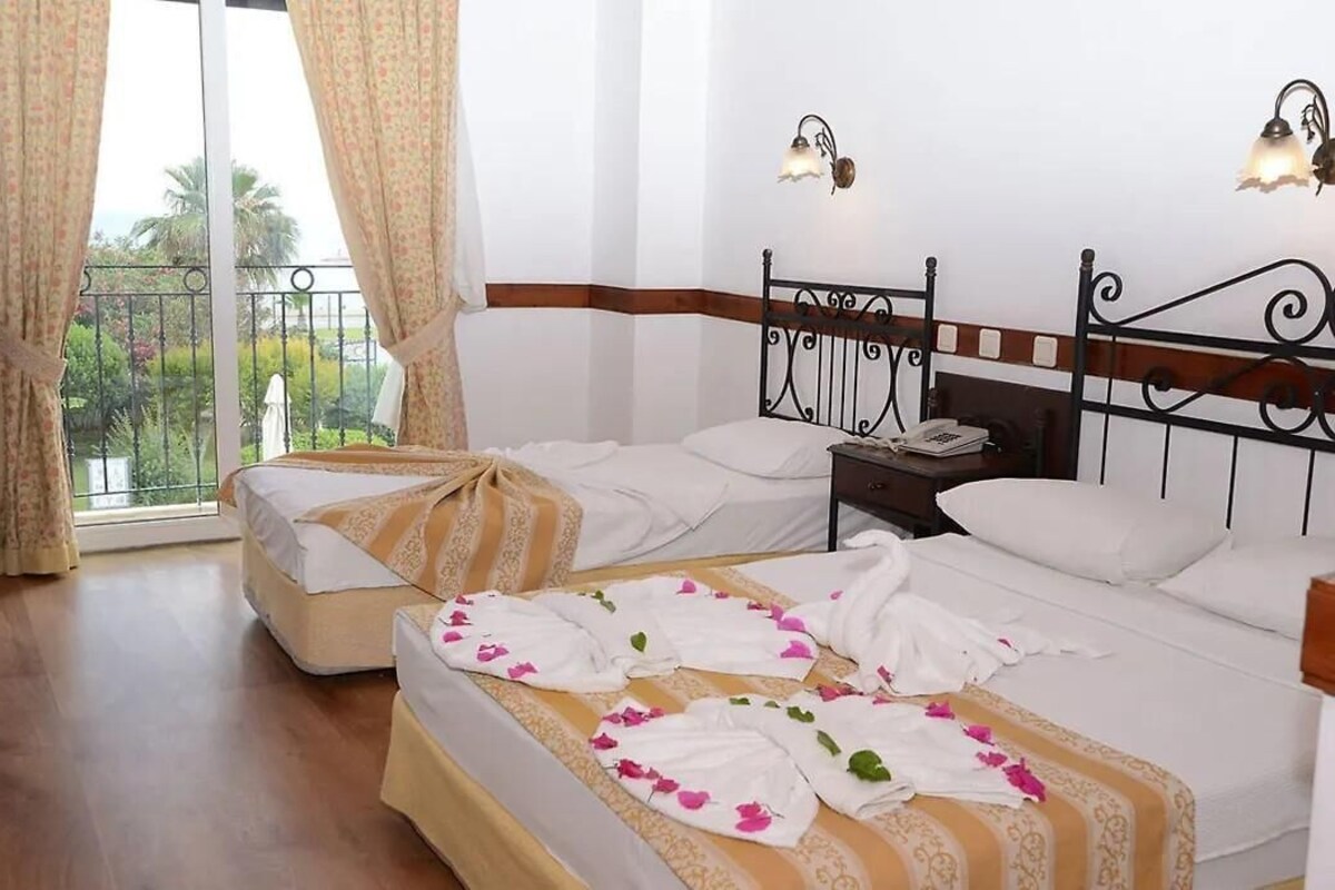 A hotel room with a balcony, a bed with white linens, a bedside table with a lamp, and a window with curtains.
