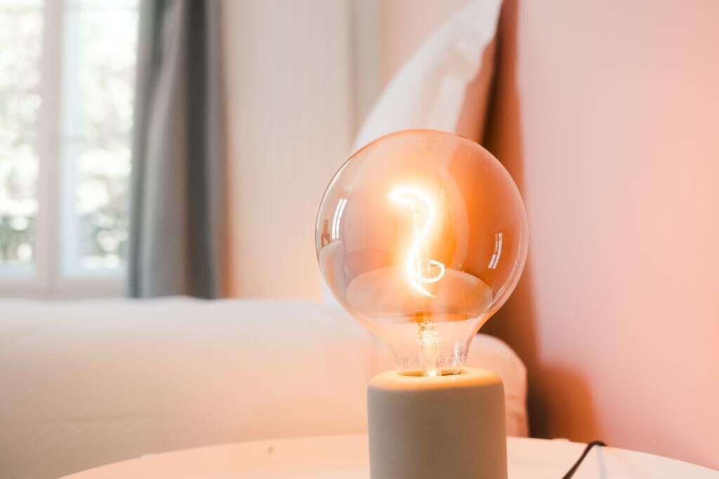 Eine klare Glühbirne auf einem weißen Sockel, vermutlich eine Tischlampe, in einem Raum mit Fenster und weißer Wand.