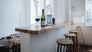 A kitchen with a wooden bar counter, two wine glasses, and a wine bottle. There are three stools and a window with a view of buildings. - La Maison sur la Loire-A stone's throw from the heart of Tours (Tours)