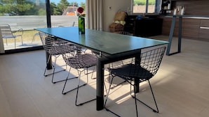 A modern dining area with a glass table and wireframe chairs, a vase with a red flower, and a view of the outdoors through large windows.