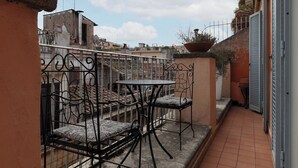 Egy kis, szabadtéri terasz, melyben egy fém asztal és székek találhatók, a padló csempézett, és kilátás nyílik a szomszédos épületekre.