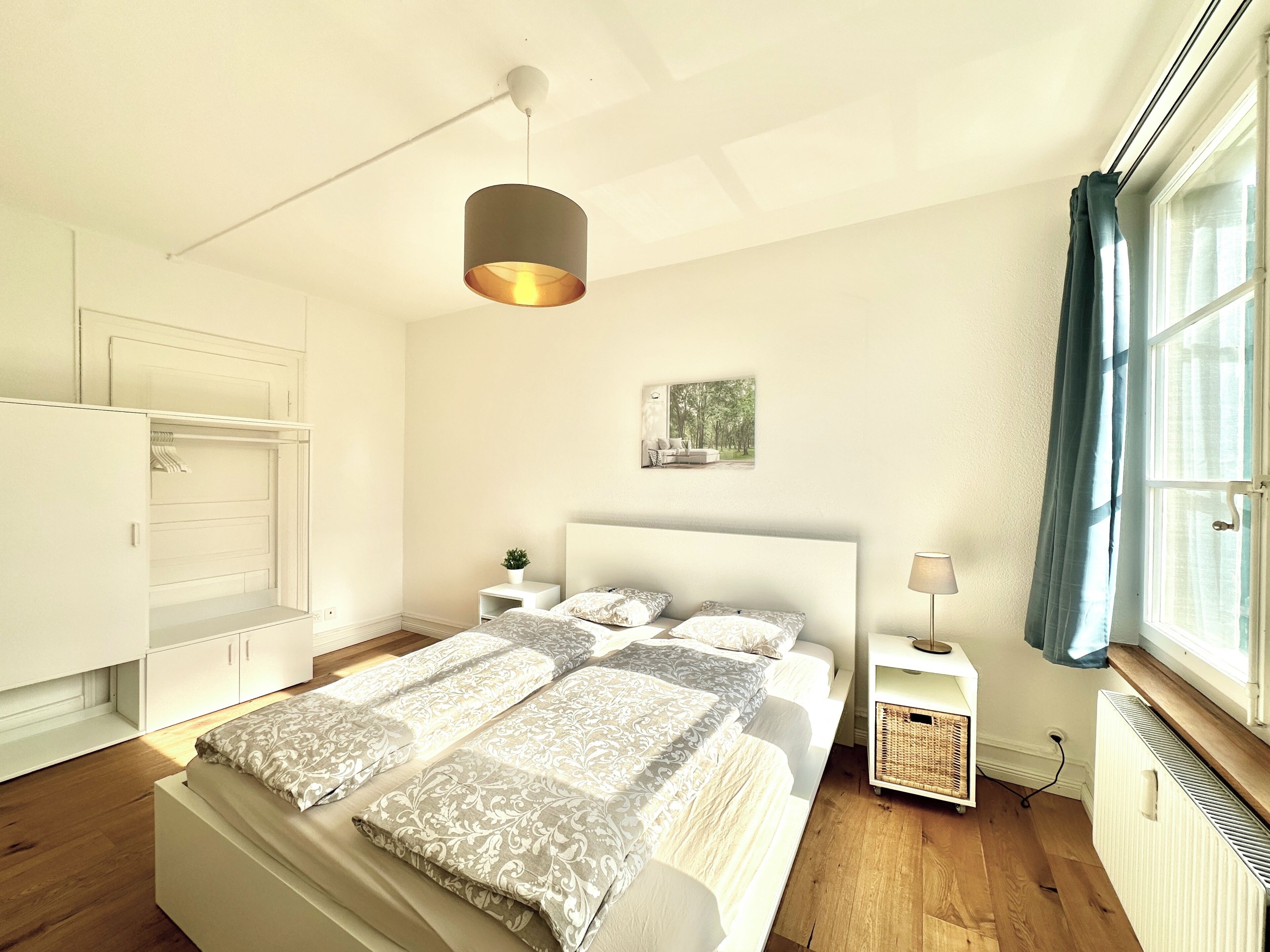 Une chambre à coucher comprenant un lit, des tables de chevet, un luminaire suspendu, une fenêtre avec des rideaux et un radiateur.