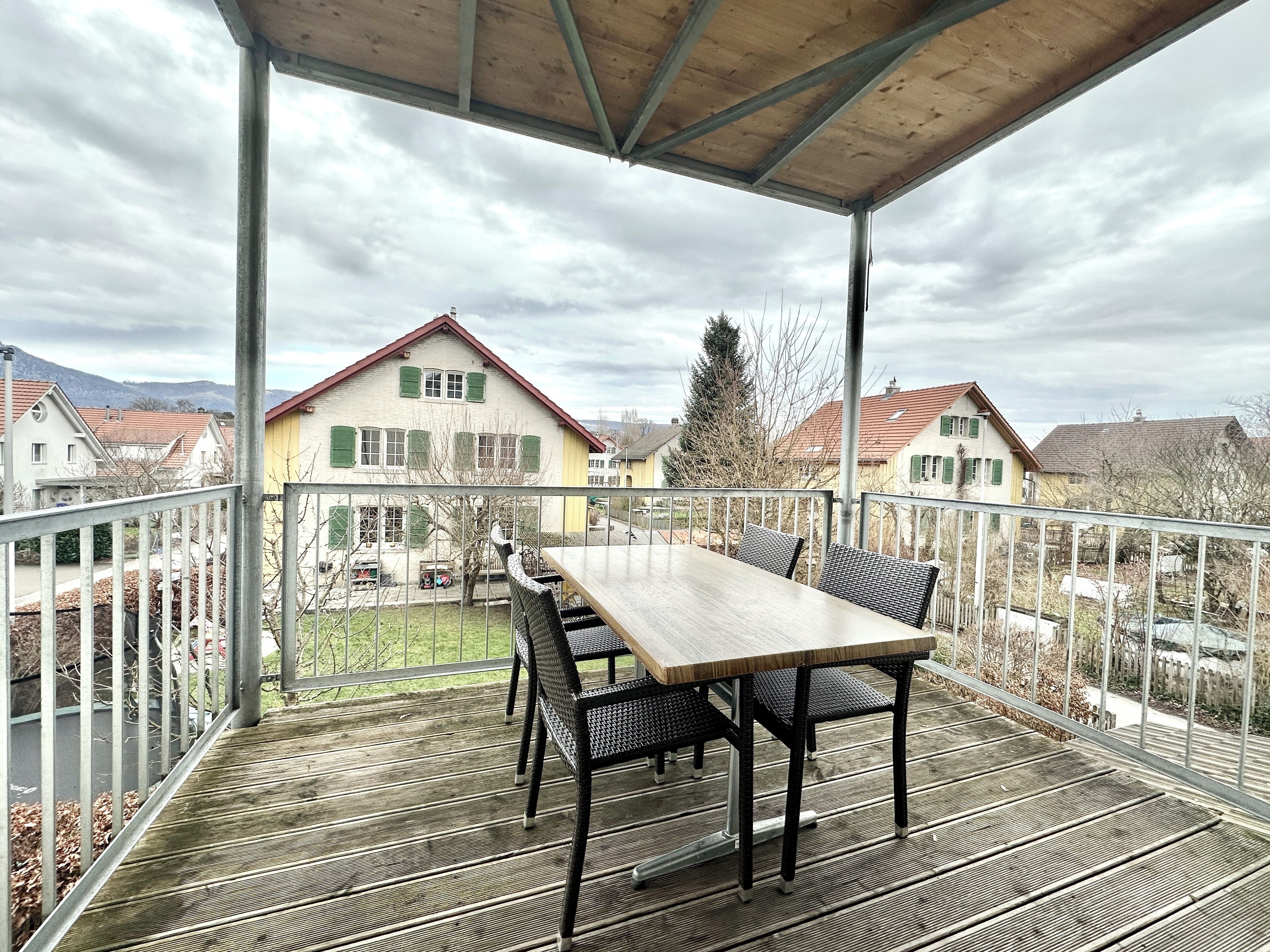 A covered outdoor area with a table and chairs, overlooking a residential neighborhood with houses and mountains in the background.