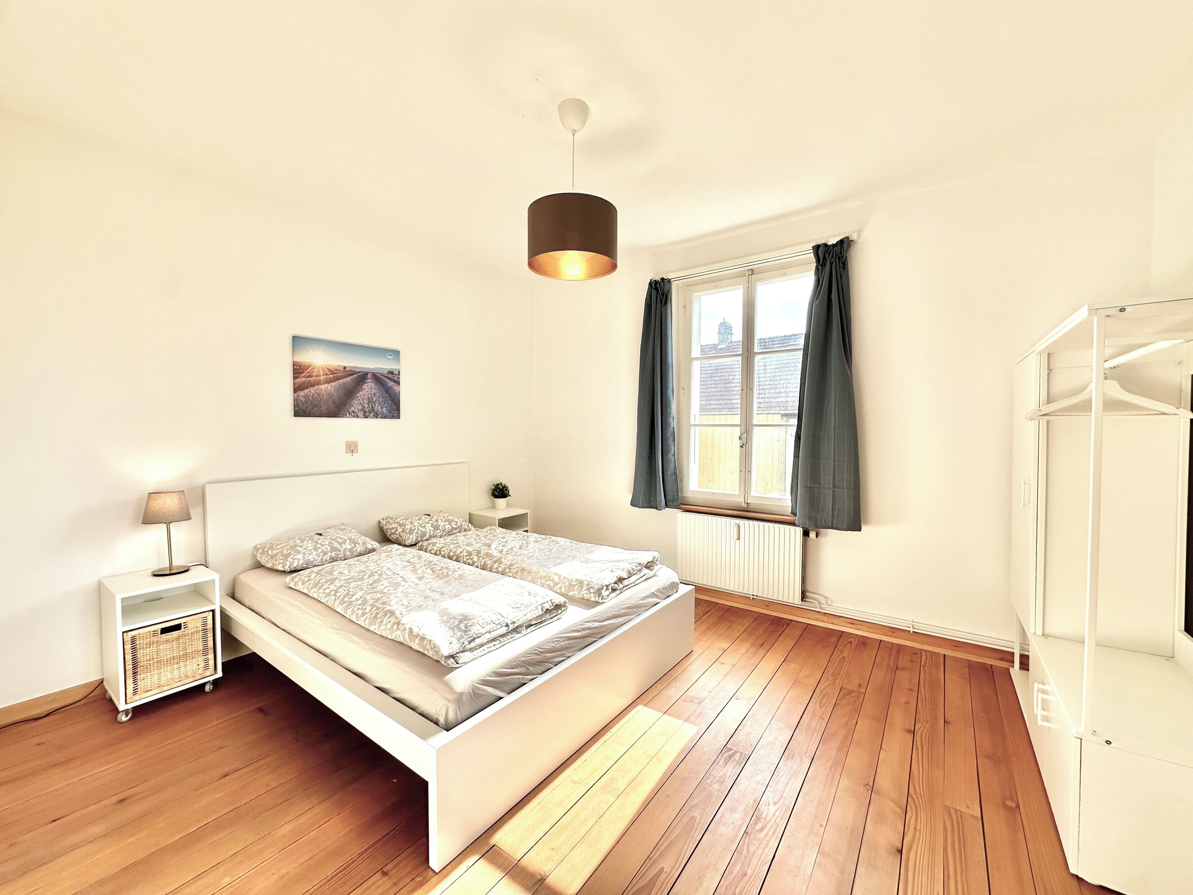 A bedroom with a double bed, a bedside table with a lamp, a window with curtains, a radiator, and a hanging light fixture.