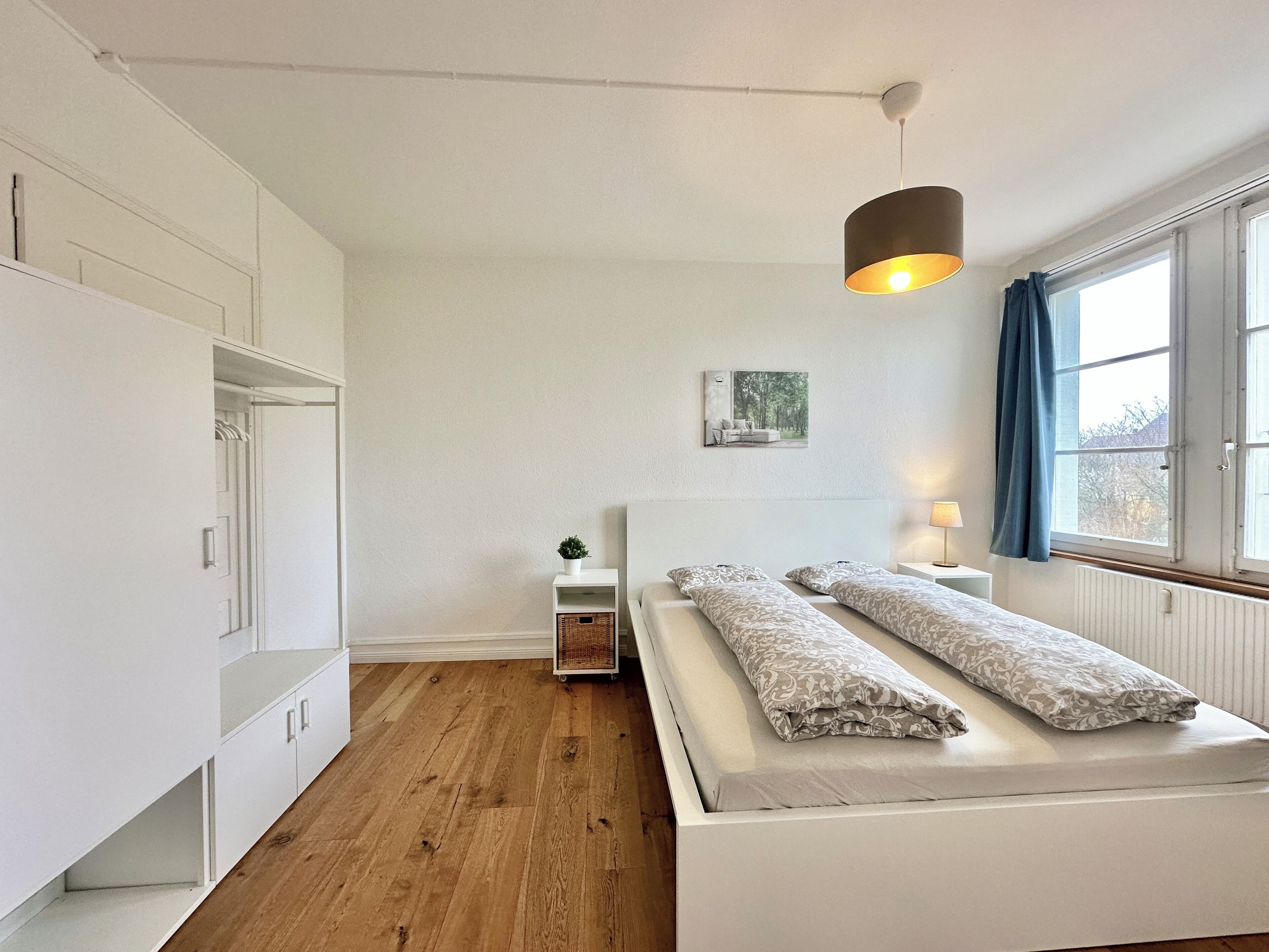 Ein Schlafzimmer mit Bett, Nachttisch, Pendelleuchte, einem Fenster mit Vorhängen und einem Wandbild.