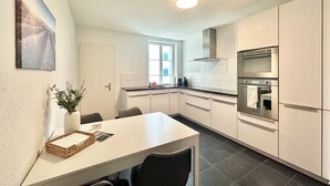 A modern kitchen with white cabinets, a dining table with chairs, a built-in oven, and a window providing natural light. - Business Apartment Derendingen (Derendingen)