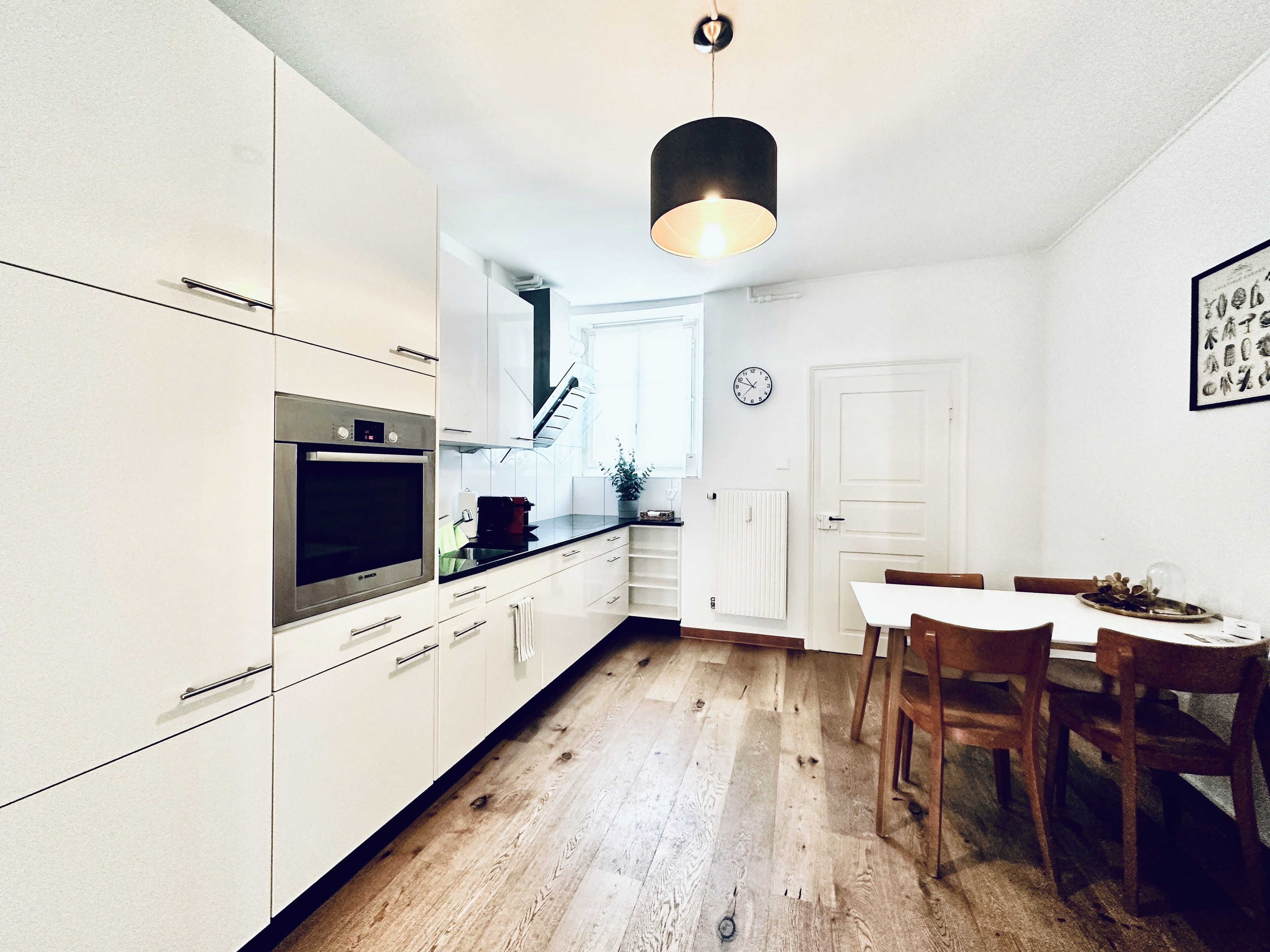 Een moderne keuken met witte kasten, een inbouwoven en een eethoek met een houten tafel en stoelen.