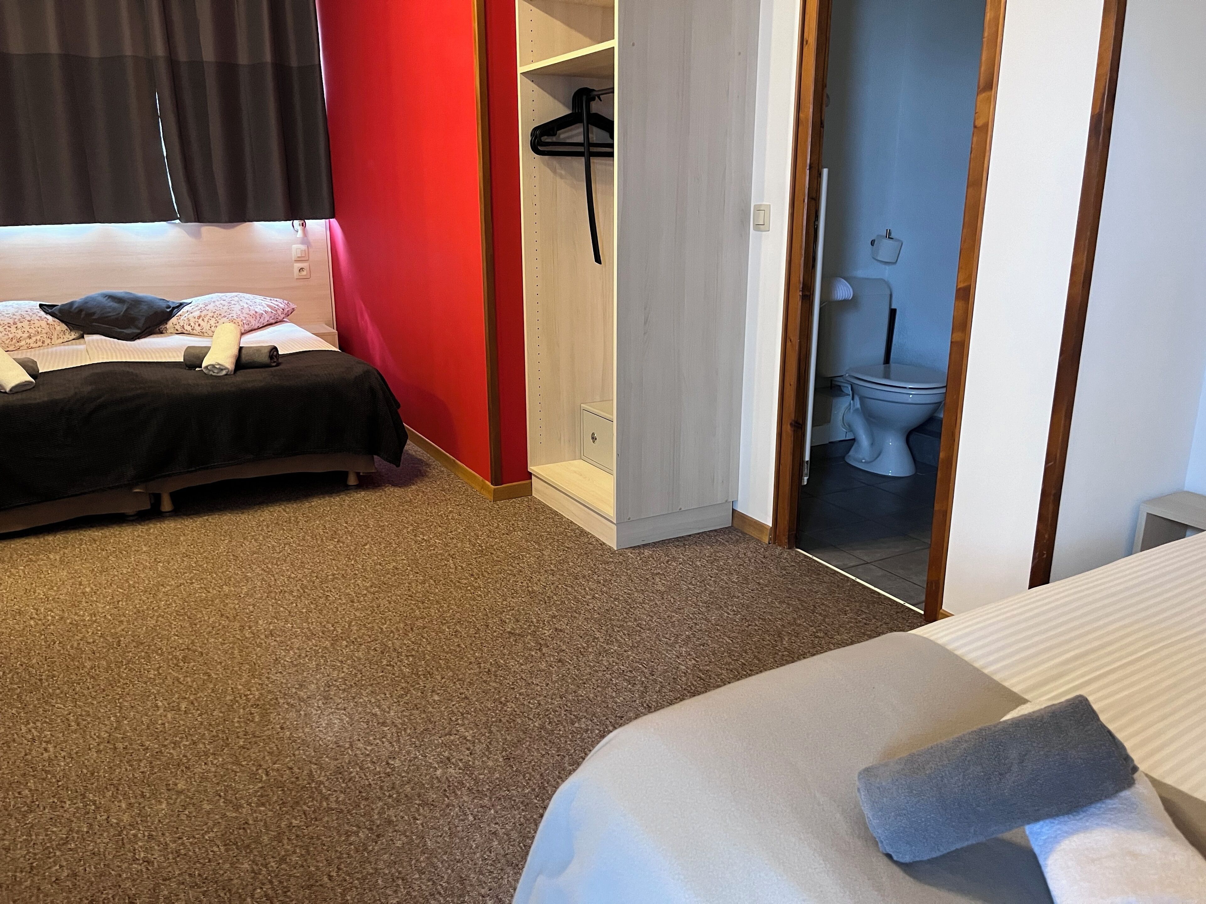 Une chambre d’hôtel avec un lit, un mur rouge, une salle de bain avec des toilettes et un lavabo, et un porte-manteau.