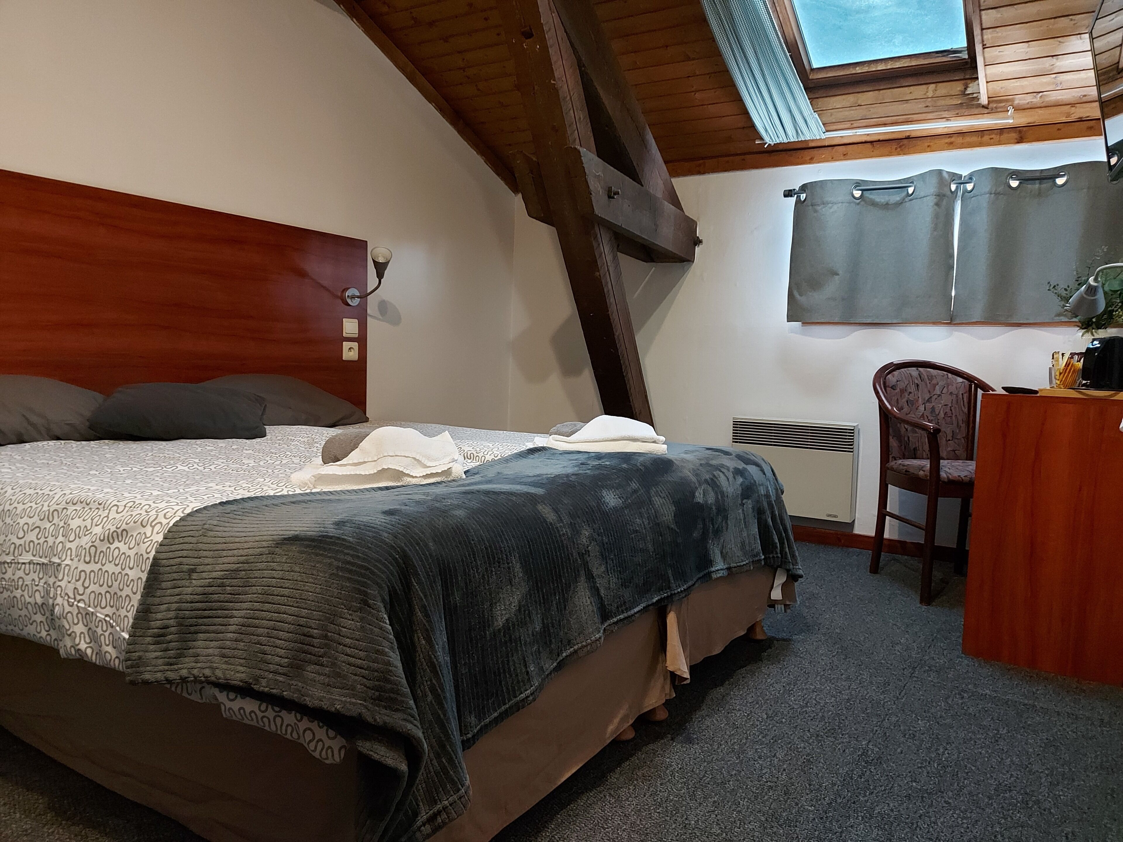 A bedroom with a wooden headboard, a bed with a patterned bedspread, a desk with a chair, and a window with curtains.
