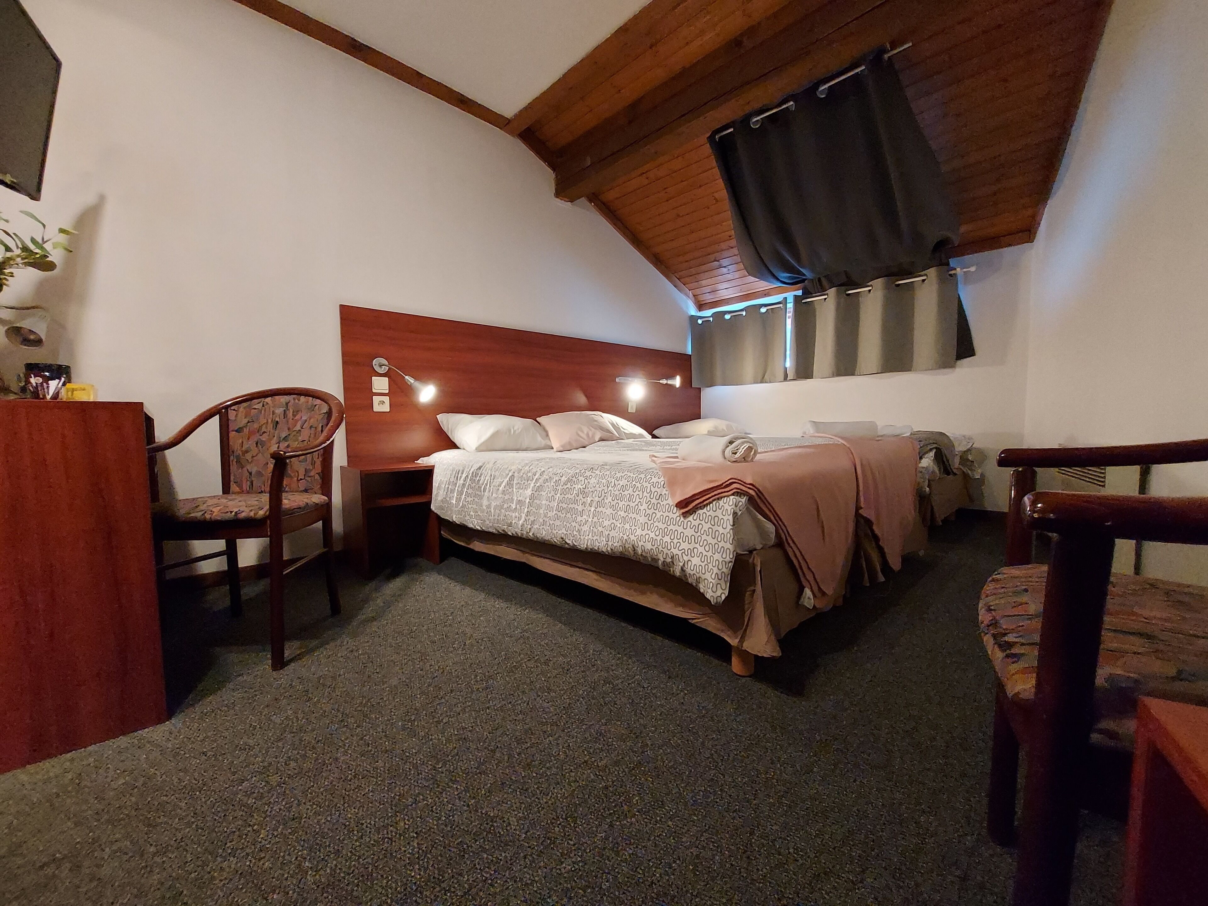 Un dormitorio con cabecera de madera, cama con edredón estampado, mesita de noche con lámpara y una silla.