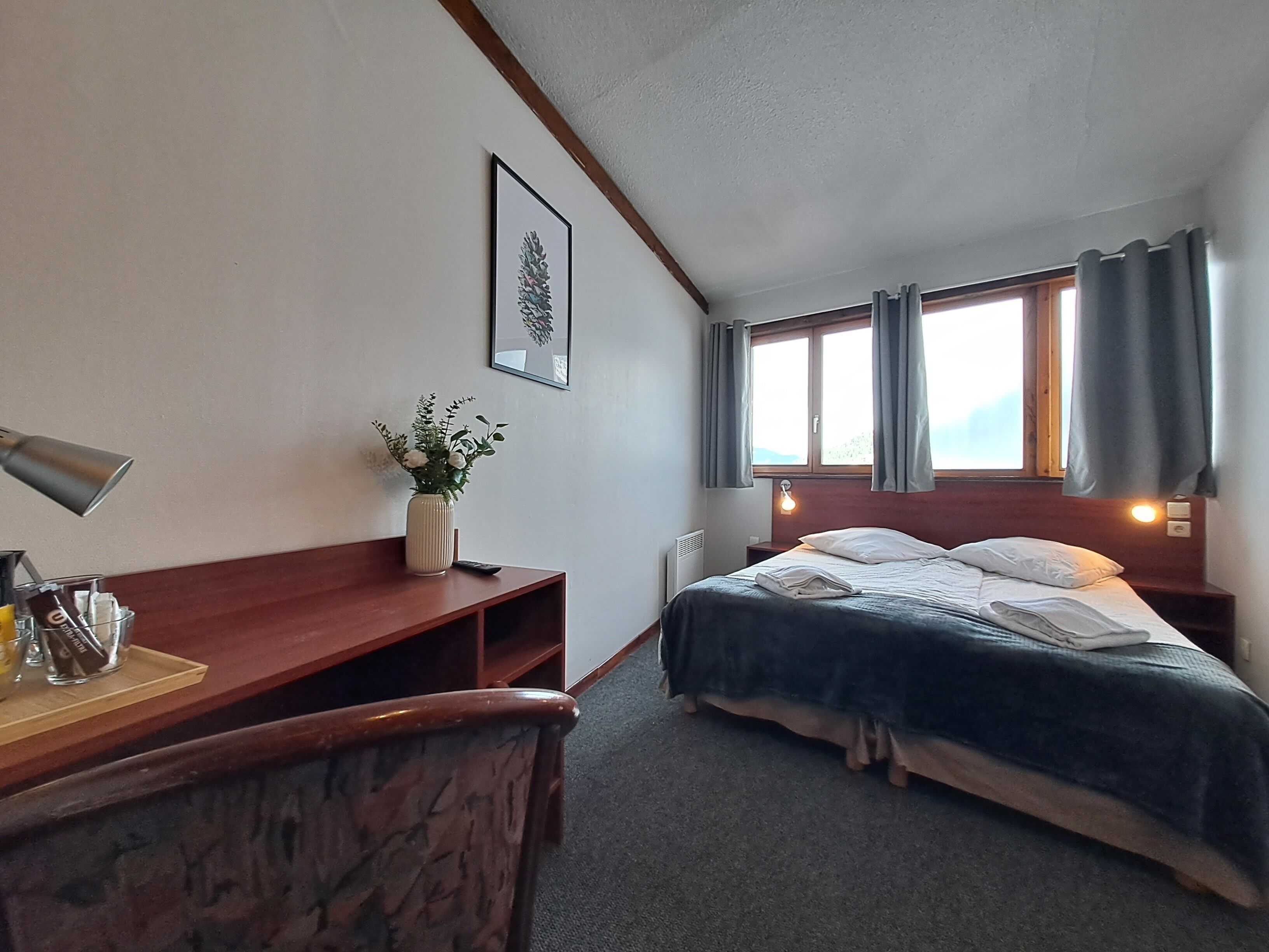 Habitació d'hotel amb llit, escriptori amb làmpada, gerra amb flors i finestra amb cortines.