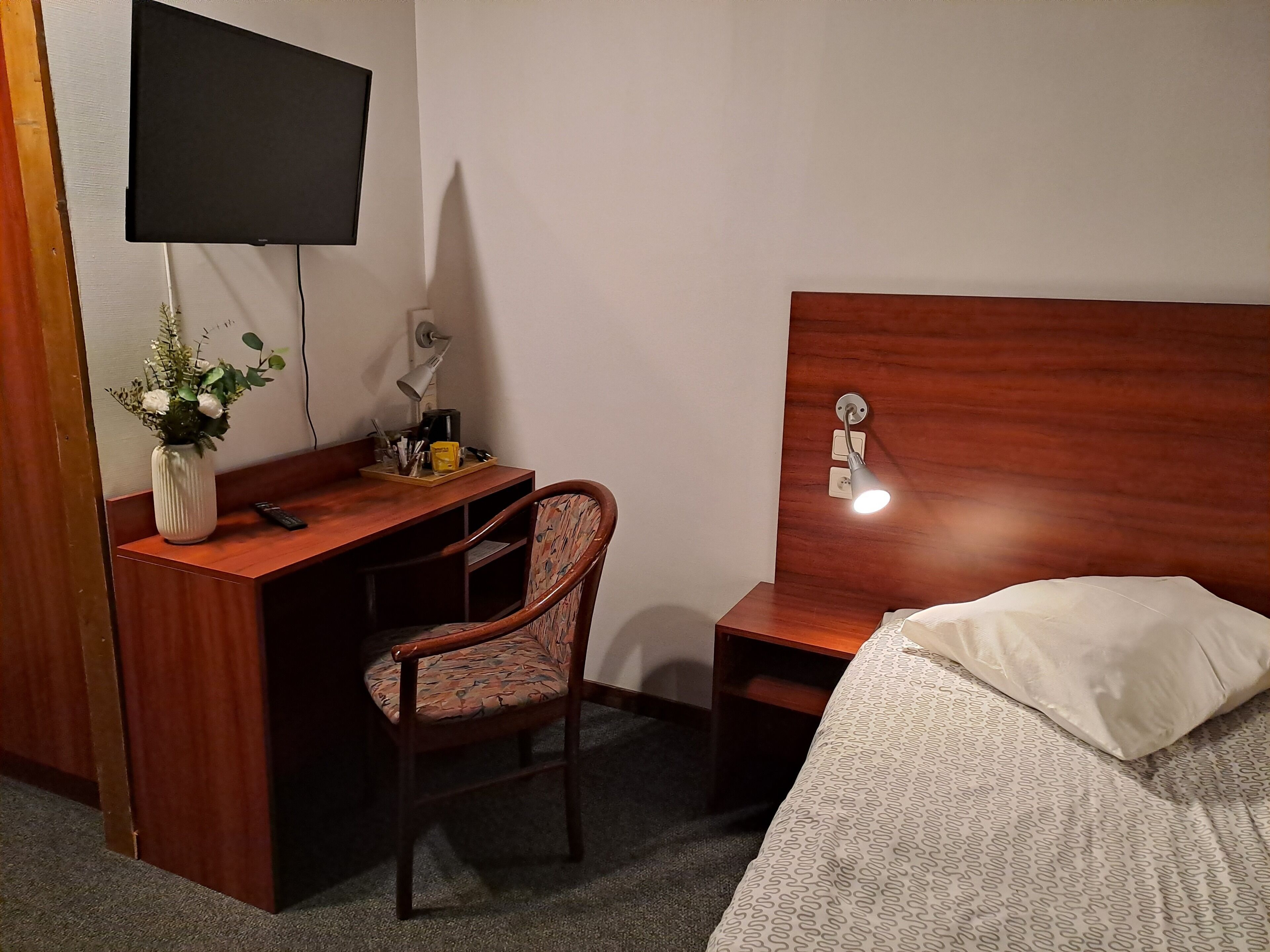 A hotel room with a wooden desk, a chair, a bed with a patterned blanket, a TV mounted on the wall, and a bedside lamp.