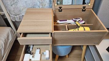 A wooden bedside table with a mirror, a drawer with electronic devices, and a stool with a cushion.