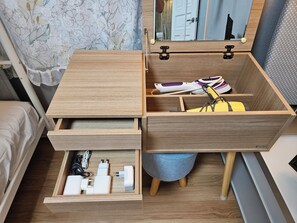 A wooden bedside table with a mirror, a drawer with electronic devices, and a stool with a cushion.