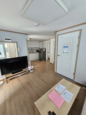 Une chambre moderne dotée d’un téléviseur à écran plat, de parquet, d’une petite table avec des documents, d’un coin cuisine et d’une porte sur laquelle est affiché un avis.