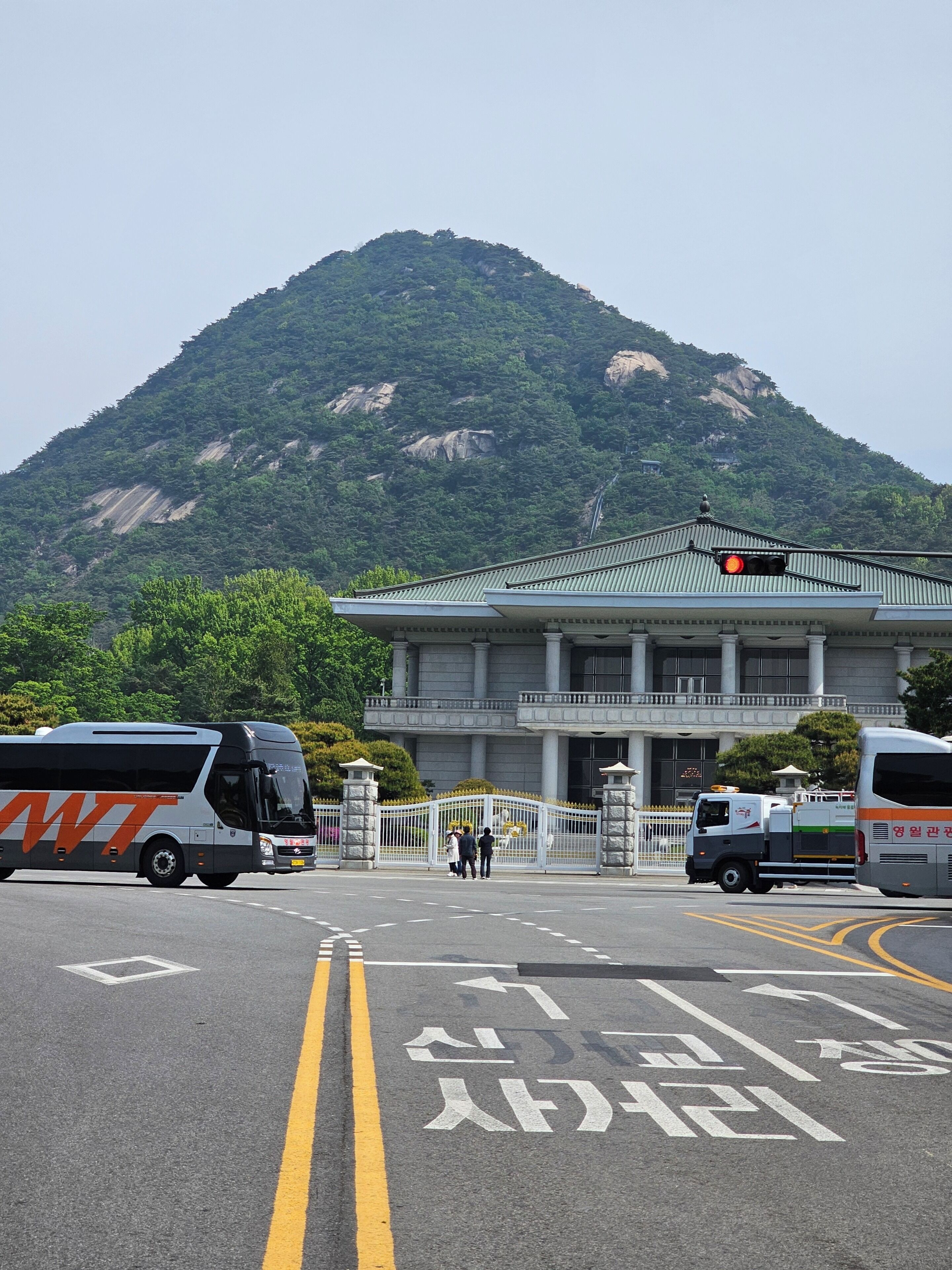 A parking lot with buses, a building with columns, and a mountain in the background.