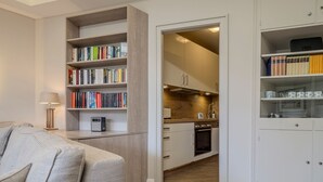 A modern living room with a sofa, a bookshelf, and a kitchen area with cabinets and appliances. - Kamperhof, App. 5 - Modern vacation apartment with 3 rooms on approx. 87 m², for max. (Sylt)