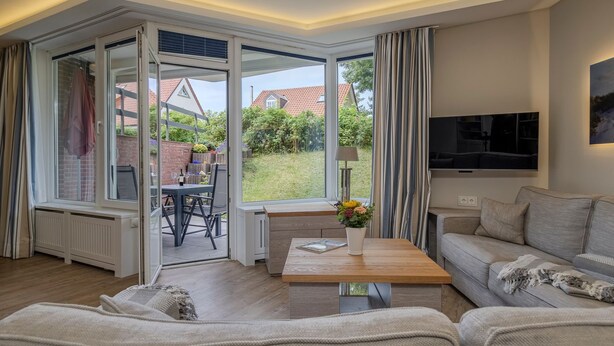 A modern living room with a sofa, a coffee table, and a TV. There is a glass door leading to an outdoor area with a table and chairs. - Kamperhof, App. 5 - Modern vacation apartment with 3 rooms on approx. 87 m², for max. (Sylt)
