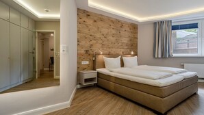 A modern bedroom with a wooden headboard, a large bed, bedside lamps, and a view of the outdoors through a window. - Kamperhof, App. 5 - Modern vacation apartment with 3 rooms on approx. 87 m², for max. (Sylt)