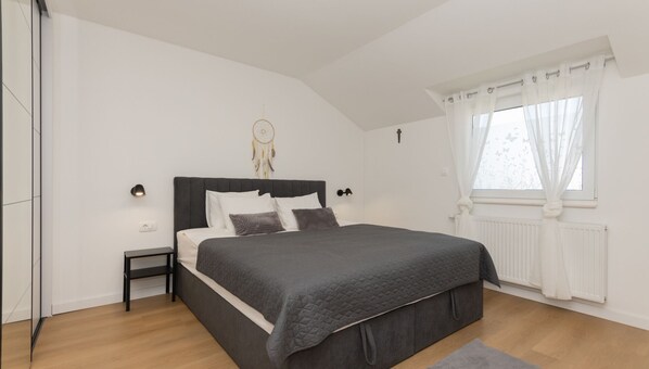 A bedroom with a large bed, a bedside table, a wall-mounted dreamcatcher, and a window with curtains.