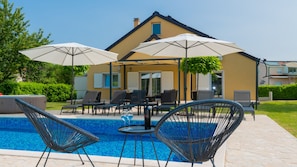 Un área de piscina con sillones de descanso y sombrillas, una casa con fachada amarilla y un cielo azul despejado.