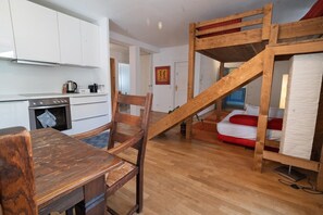 A compact room with a wooden table, a chair, a bunk bed, a kitchen area with cabinets and appliances, and a staircase.