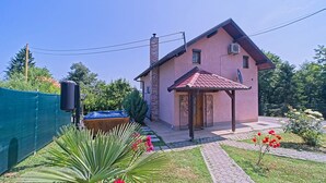 Una casa rosa con techo de tejas, un jacuzzi de madera y un jardín con flores rojas y plantas verdes.