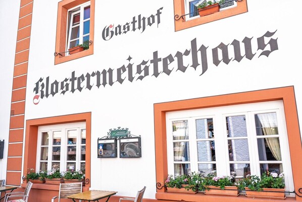 A building with a sign reading 'Gasthof Klostermeisterhaus' and outdoor seating with flower pots.