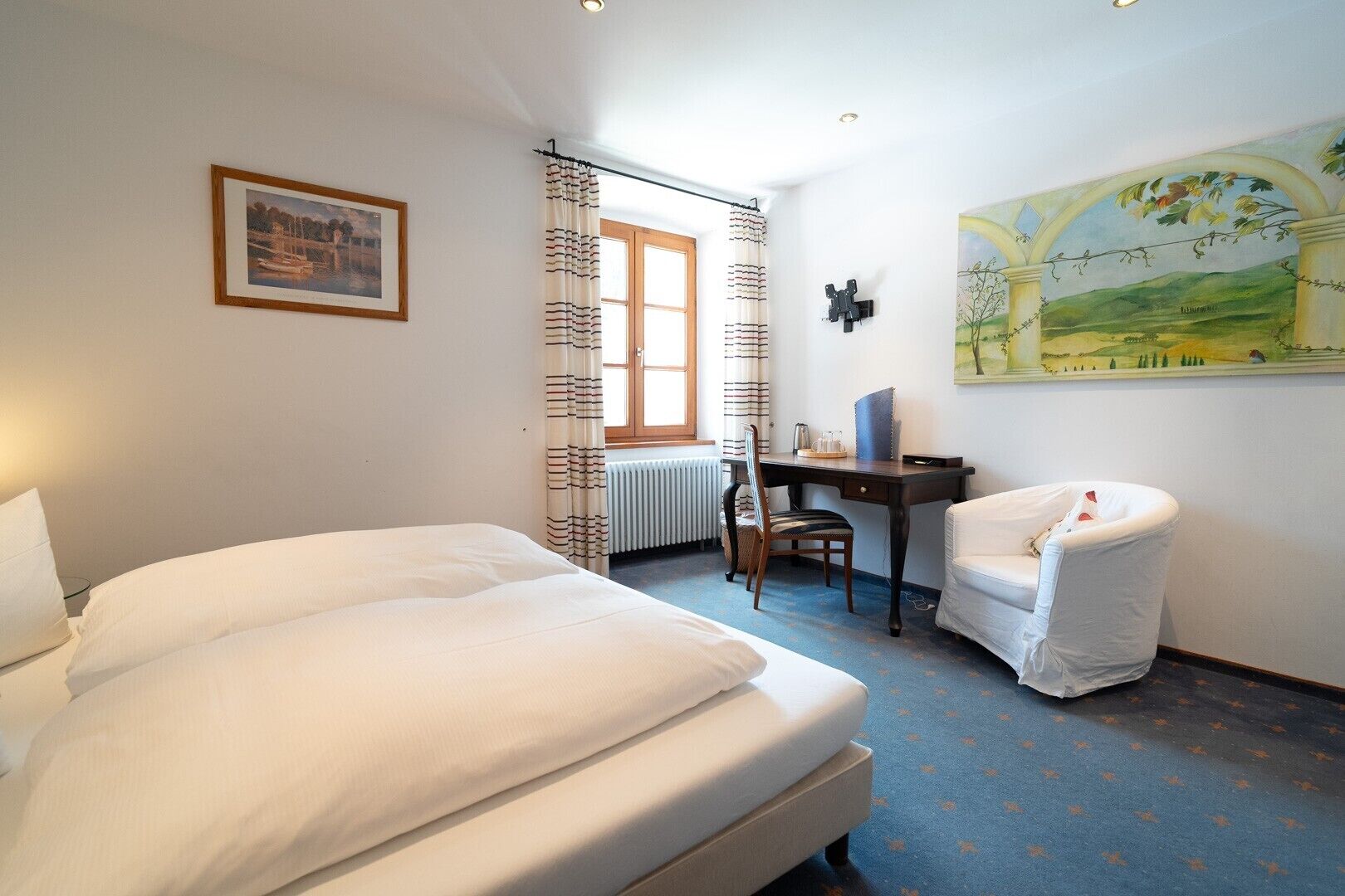 A hotel room with a bed, a desk, a chair, and a painting on the wall.