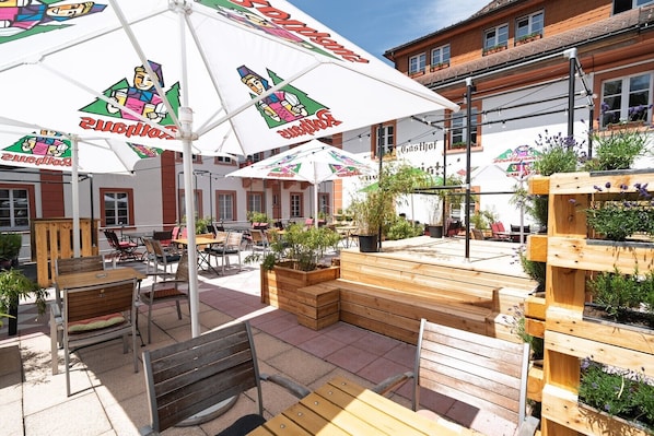 An outdoor seating area with wooden benches and tables, umbrellas, and potted plants.
