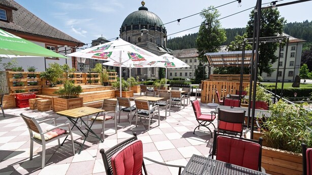 Ein Terrassenbereich im Freien mit Tischen und Stühlen, einem großen Sonnenschirm und einem Gebäude mit Kuppel im Hintergrund.