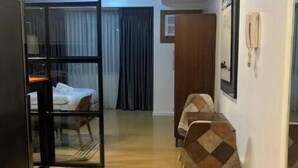 A hotel room with a wooden floor, a bed with a white cover, a dining area with a table and chairs, and a door leading to another room. - Cozy studio unit in Avant BGC (Taguig)