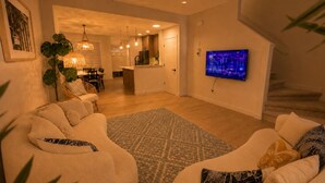 Een moderne woonkamer met een zithoek, een kamerkleed met dessin, een aan de muur bevestigde televisie, en een trap op de achtergrond.