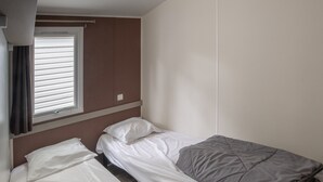 En enkeltseng med hvidt og gråt sengetøj, et vindue med rullegardiner og en brun væg.