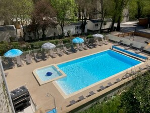 En rektangulär swimmingpool med ett bubbelbad, omgiven av solstolar och parasoller, belägen i ett vackert utformat område med träd och ett staket.