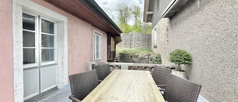 An outdoor patio with a wooden table and wicker chairs, a stone wall, and a wooden fence in the background.