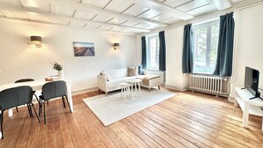 A modern living room with a white sofa, a dining table with chairs, a TV, and a large window with curtains. - Business Apartment Oberbipp (Oberbipp)