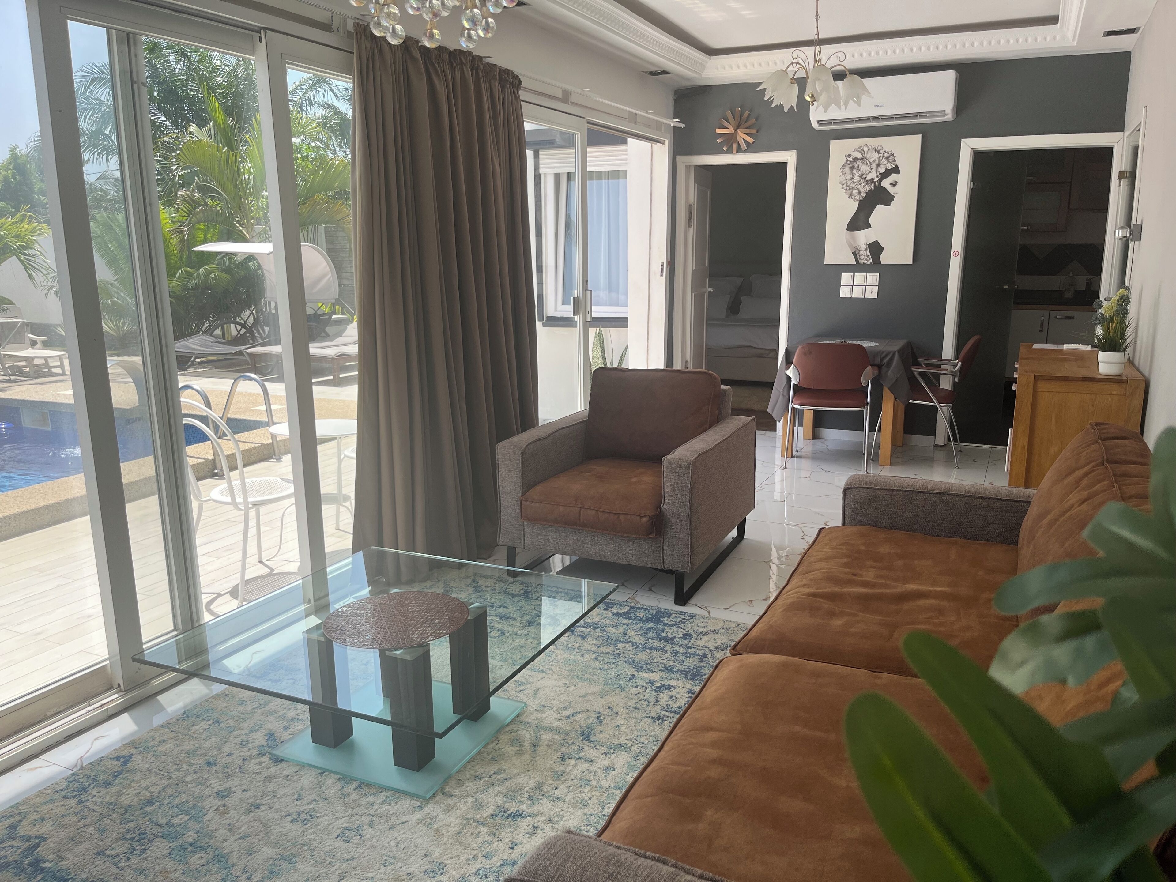 A spacious living room with a glass coffee table, a sofa, and a chair. A large window offers a view of a pool and palm trees.