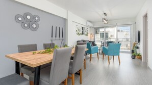 A modern dining area with a wooden table, grey upholstered chairs, and a blue chair. There is a wall art piece and a potted plant on the wall. - Amore 2-BR Apt By Finikoudes (Larnaca)