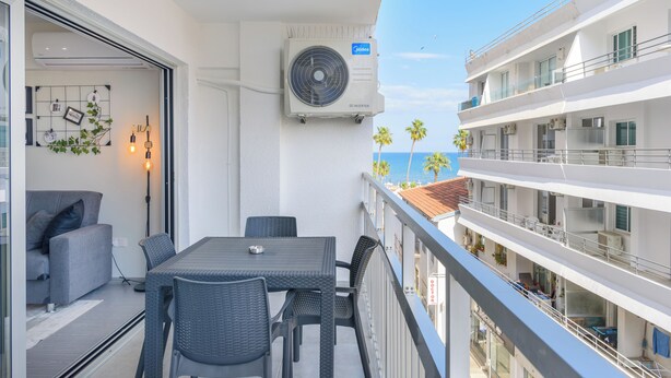 A balcony with a table and chairs, a fan, and a view of the sea and palm trees. - Amore 2-BR Apt By Finikoudes (Larnaca)