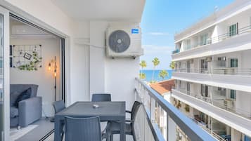 Uma varanda com mesa e cadeiras, um ventilador e vista para o mar e palmeiras.