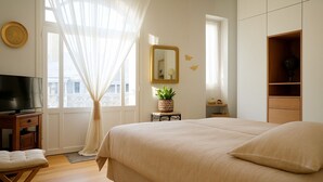 Une chambre avec un grand lit, un téléviseur posé sur un meuble en bois, une fenêtre avec des rideaux légers et une petite table sur laquelle se trouve une plante.