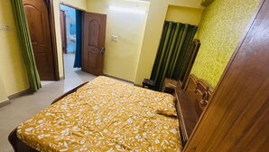 A bedroom with a bed, a wooden wardrobe, a small stool, and a door leading to another room. - Prashi Homes (Varanasi)