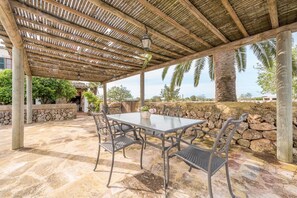 Een terras buiten met een tafel en stoelen, een pergola erboven en een stenen muur.